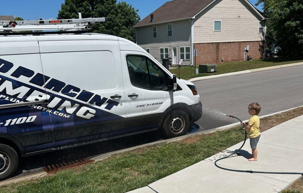 A 100 Percent Plumbing service van being washed on a Nashville-area street, with a small child helping by holding the hose